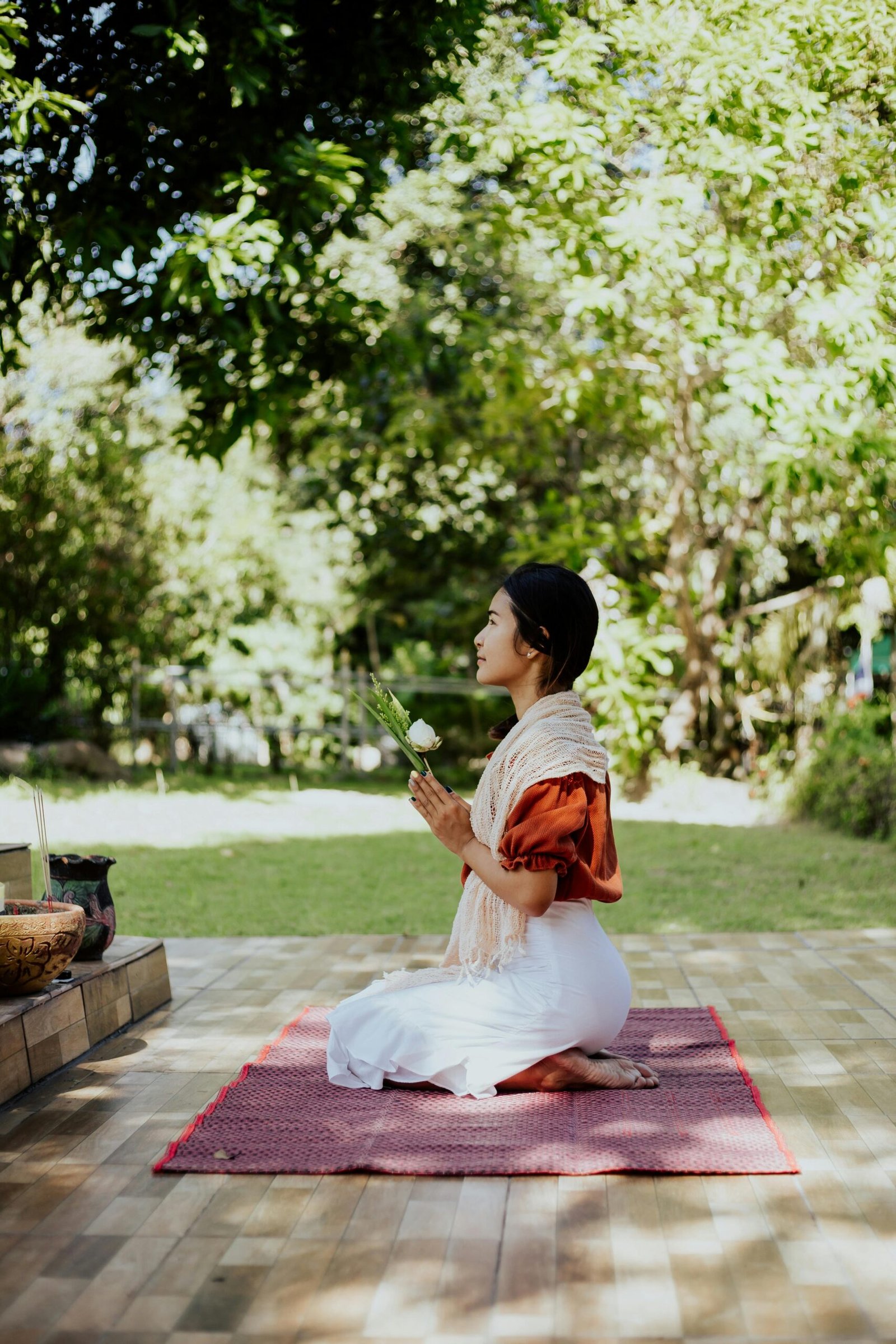 A woman kneeling outdoors, praying with a flower in a serene garden setting.